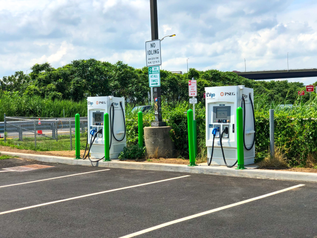 Ladestation für Elektrofahrzeuge auf einem Parkplatz mit umgebennder Infrastruktur, Grünfläche und einer Brücke im Hintergrund.