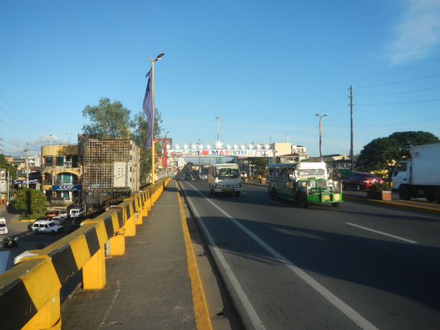Volles Stadtbild mit Fahrzeugen, einer Mitteltrennung, einem Fahnenmast mit Fahne, Gebäuden, Bäumen, Strommasten mit Drähten und einem bewölkten Himmel.