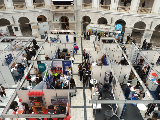 Große Gruppe von Teilnehmern auf einer internationalen Fachmesse mit Ständen, Bannern, Tischen, Stühlen, Luftballons und einem Gebäude mit Bögen, Säulen, Geländern und einem Bildschirm im Hintergrund.