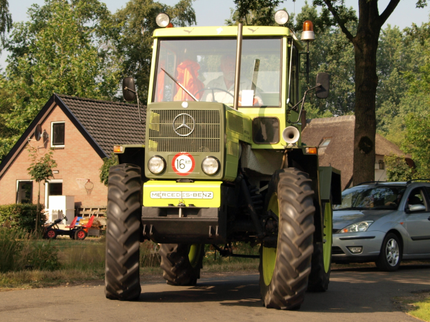 Ein grüner Traktor fährt auf einer Straße neben einem Auto, mit einer Person auf dem Fahrersitz des Traktors, vor einem Hintergrund aus Häusern, Bäumen, Pflanzen, Gras und einem klaren blauen Himmel.