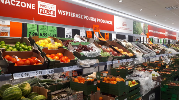 Ladeninneres mit einer Vielzahl von Obst- und Gemüsesorten in Kartonschachteln mit Preisschildern, beleuchtet von Deckenlampen und mit informativen Schildern an den Wänden.