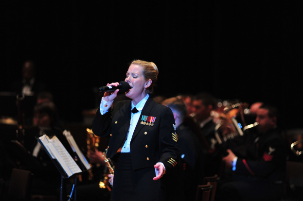 Frau in militärischer Uniform, die in ein Mikrofon singt, begleitet von einer Band, die Instrumente auf einer dunklen Bühne spielt, mit Büchern auf Ständern davor.