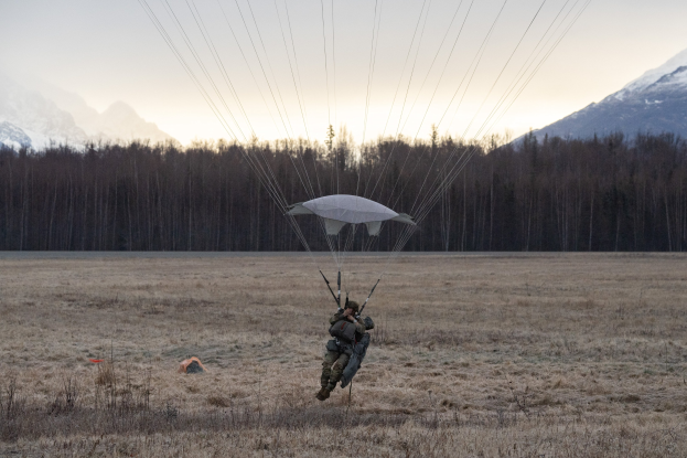 Eine Person beim Paragleiten über ein grasbewachsenes Feld mit schneebedeckten Bergen im Hintergrund, die einen Helm trägt und von grünen Bäumen umgeben ist unter einem klaren blauen Himmel.