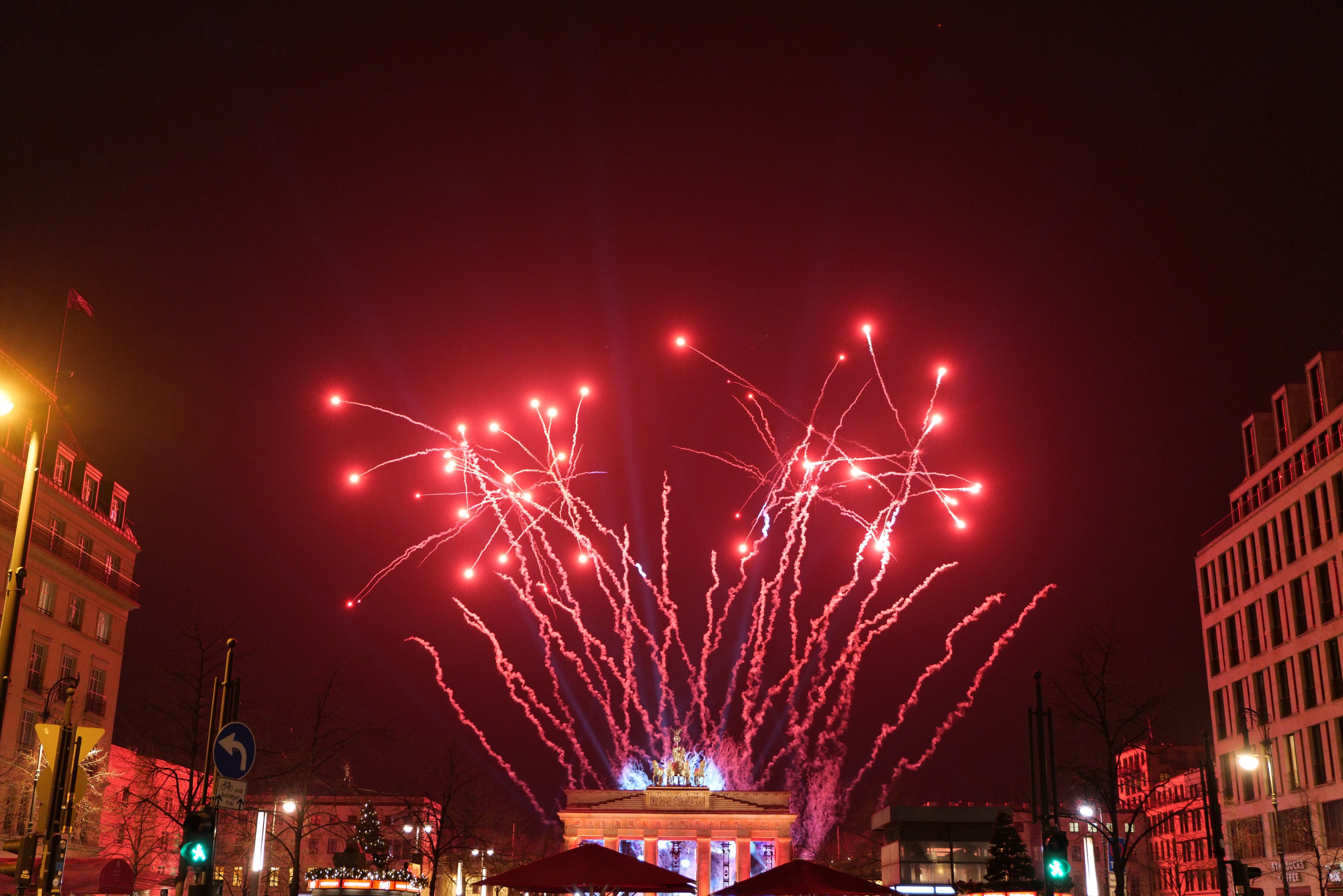 Eine belebte Stadtstraße bei Nacht am Neujahrstag in Berlin, mit Gebäuden, Bäumen, Laternen, Ampeln, Schildern, Zelten, Menschen und einem prächtigen Feuerwerk, das den Himmel erleuchtet.