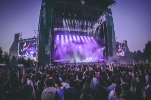 Eine große Menschenmenge auf einem Musikfestival mit einer Bühne, auf der Musiker spielen, beleuchtet von Lichtern und umgeben von Bildschirmen, vor einer Kulisse aus einem Gebäude, Bäumen und einem klaren blauen Himmel.