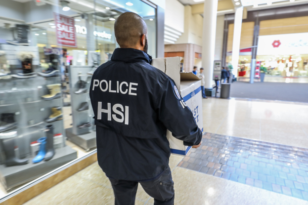 Polizeibeamter in schwarzer Jacke mit einer Kiste, der durch ein Einkaufszentrum geht, mit anderen Menschen, einem Müllcontainer, Säulen, Deckenlampen und Regalen mit Schuhen im Hintergrund.