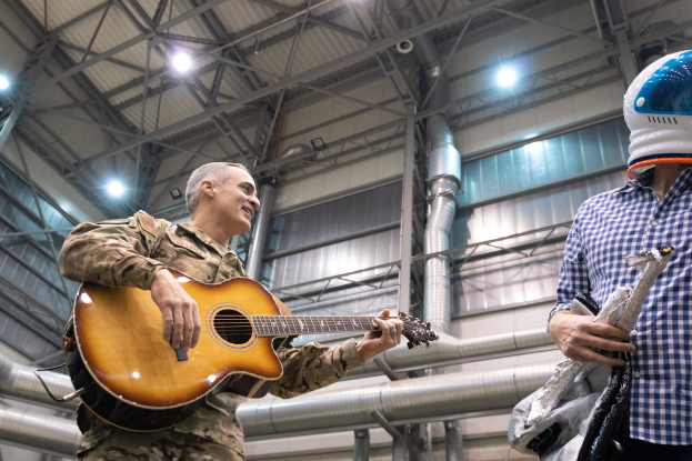 Ein Mann in einer Militäruniform spielt eine Gitarre, während er neben einem anderen Mann in einem Helm steht, mit Rohren und Lichtern im Hintergrund.