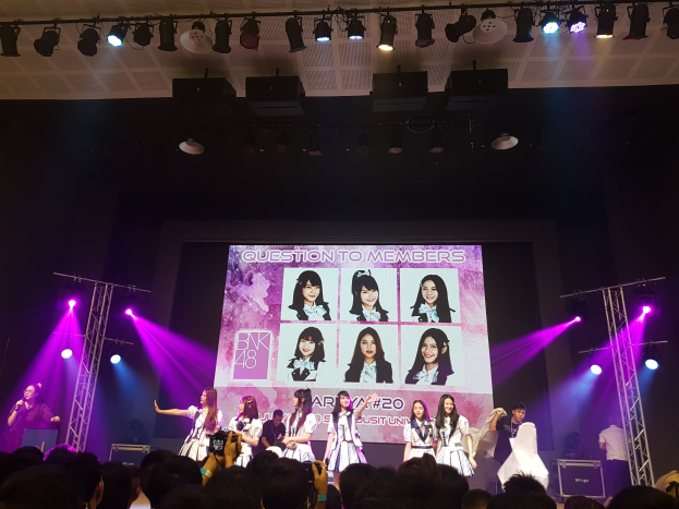 Eine Gruppe von Menschen auf einer hell erleuchteten Bühne mit Trägern, wobei einer ein Mikrofon hält und ein anderer ein Instrument spielt, während eine Menge mit Kameras unten zusieht; ein großer Bildschirm im Hintergrund zeigt Bilder der K-Pop-Gruppe BK48.