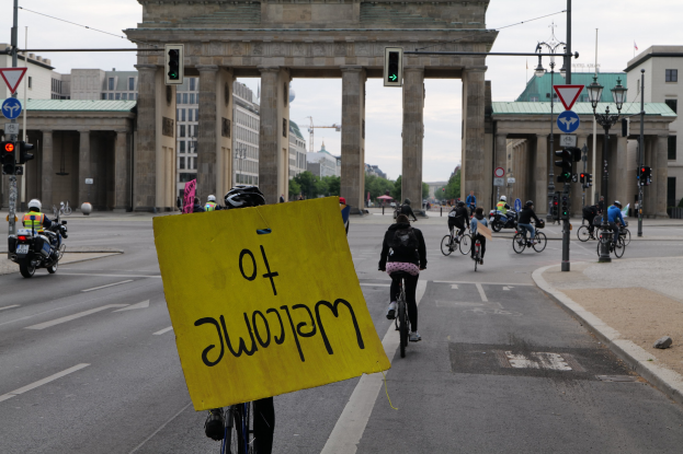 Eine Gruppe von Menschen, die auf Fahrrädern die Straße vor dem Brandenburger Tor in Berlin, Deutschland, entlangfahren, wobei eine Person ein gelbes Schild hält und der Hintergrund Gebäude, Bäume und einen klaren blauen Himmel zeigt.