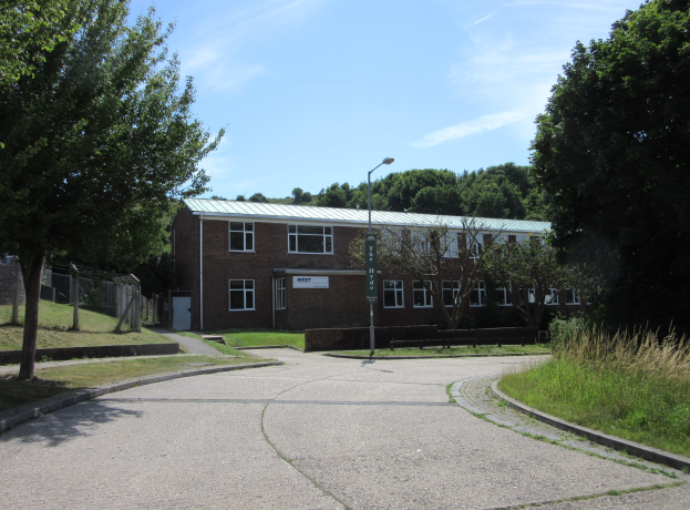 Ein Schulgebäude mit Fenstern, ein Straßenpfahl, ein Schild, eine Baumgruppe, Gras, ein Zaun und ein bewölkter Himmel, umgeben von einem Park mit grünem Gras.