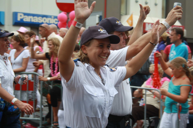 Eine Gruppe von Menschen, einige mit Mützen und Dosen tragend, steht und sitzt vor einer Menge, mit Geländern, Luftballons und einem Gebäude mit Namensschildern im Hintergrund.