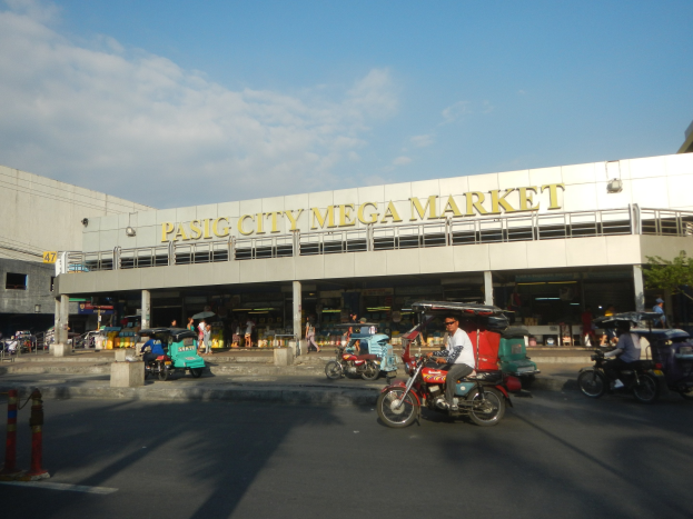 Eine belebte Straßenszene am Pasig City Mega Market mit Fahrzeugen und Fußgängern, mit einem Gebäude mit Text im Hintergrund unter einem bewölkten Himmel.