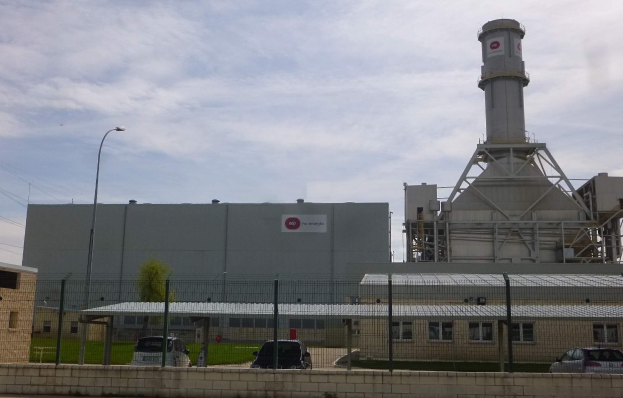 Ein großes Gebäude mit einem zentralen Turm, umgeben von einem Zaun, mit Fahrzeugen auf der benachbarten Straße, Masten, Lampen, Drähten, Bäumen und Gras im Vordergrund und einem bewölkten Himmel im Hintergrund, Teil einer neuen Energiespeicheranlage.