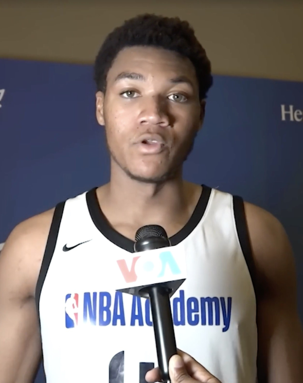 Ein Mann in einem weißen Tanktop hält ein Mikrofon und spricht vor einem Banner mit der Aufschrift "NBA All-Star Game 2019" und einer Wand.