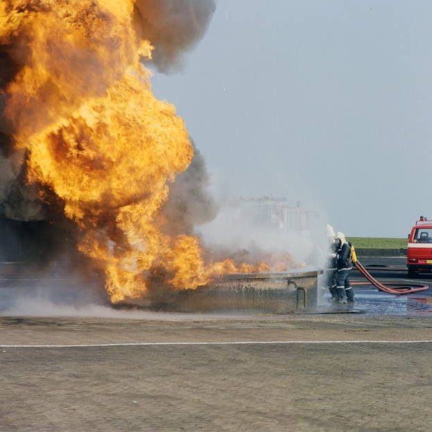 Feuerwehrlöschfahrzeug von Flammen eingehüllt auf der Seite einer Straße mit zwei Helikopterpiloten, die Rohre halten, ein Fahrzeug im Hintergrund und der Himmel sichtbar.