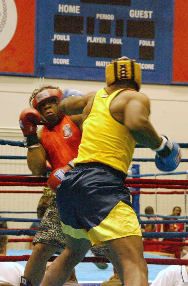 Zwei Boxer im Ring, umgeben von einer Menge, mit einer Tafel im Hintergrund, die Text und Logos anzeigt.