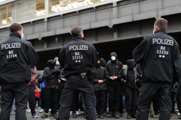 Polizisten in schwarzen Uniformen und Masken stehen vor einer Menge auf einer Demonstration, mit einer Brücke und einem Gebäude im Hintergrund.