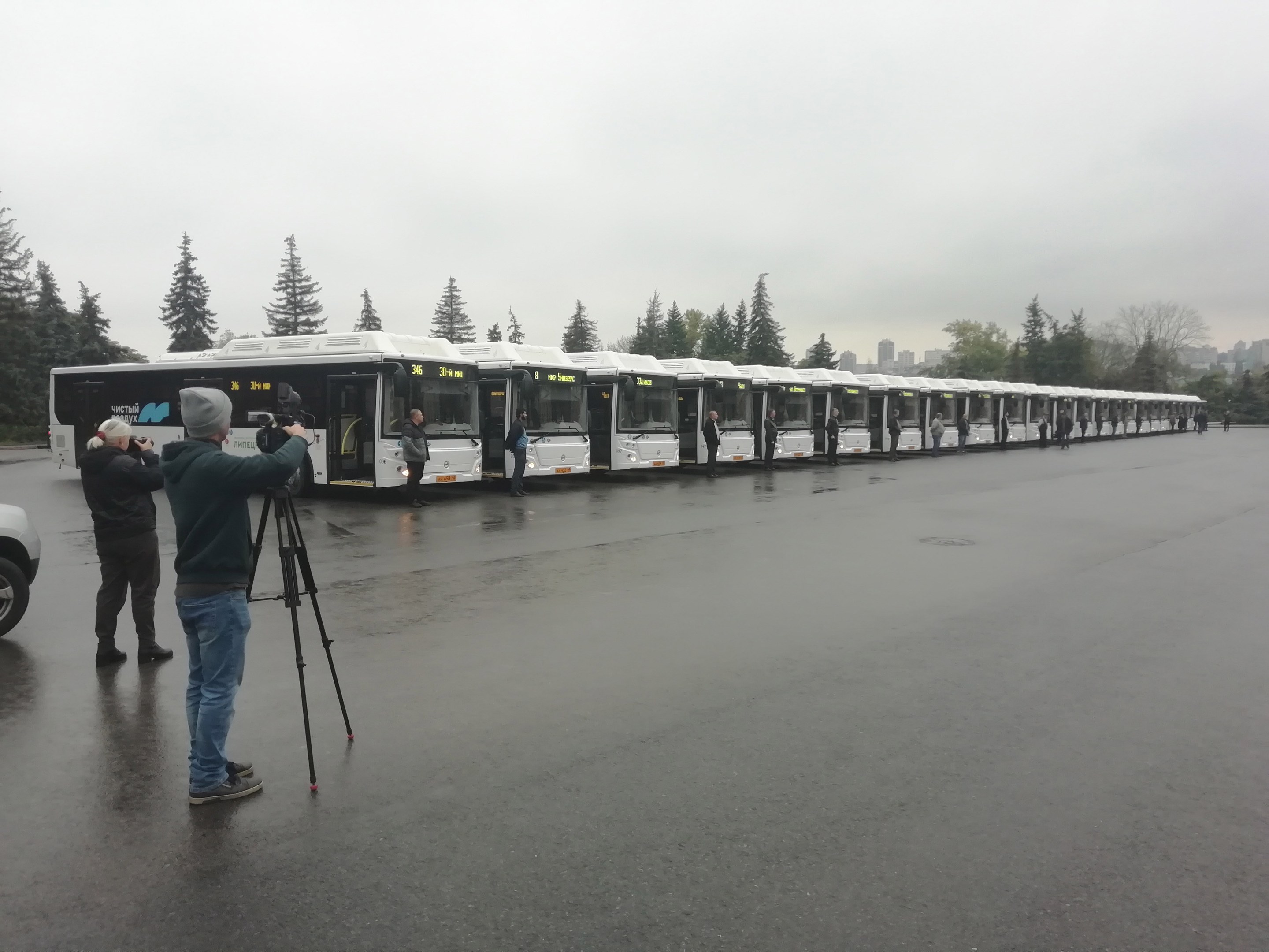Eine Gruppe von Menschen, die Fotos von einer Reihe von Bussen auf einer Straße mit Bäumen, Gebäuden und einem klaren blauen Himmel im Hintergrund machen.