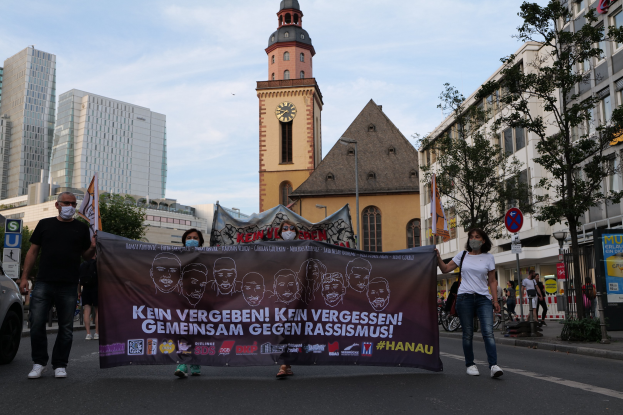 Eine Gruppe von Menschen in Masken geht eine Straße entlang, hält ein Banner mit Text und Bildern, mit einem geparkten Auto auf der linken Seite, Gebäuden, Bäumen, Schildern, Pfosten, einem Uhrenturm und einem klaren blauen Himmel im Hintergrund.