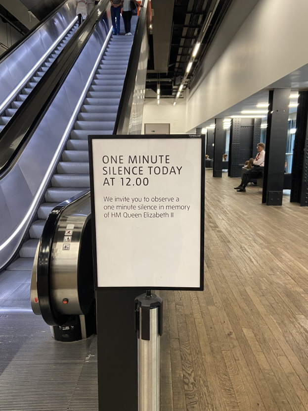 Eine Rolltreppe in einem Flughafen mit einem Schild, auf dem "Eine Minute Stille heute" steht, sowie ein paar Menschen darauf und an der Decke befestigte Lichter im Hintergrund.