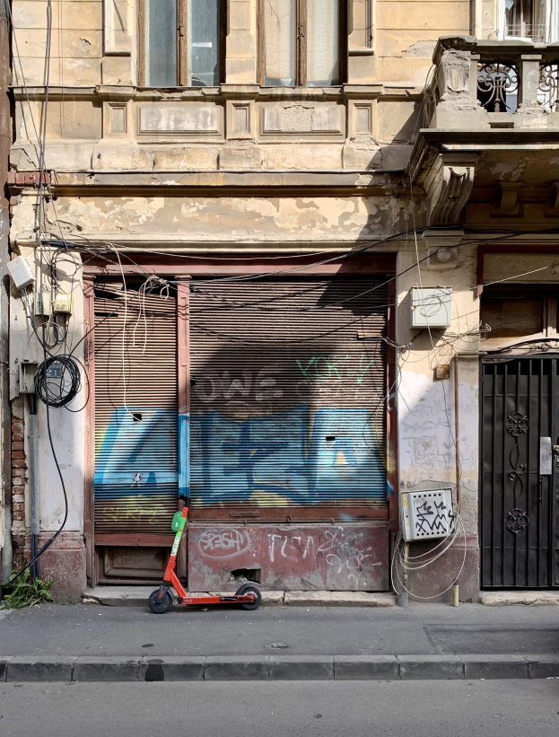 Ein roter Roller, der auf einer Straße neben einem Gebäude mit Fenstern, Rollläden und einem Tor geparkt ist, mit sichtbaren Kabeln und anderen Gegenständen entlang der Straße.