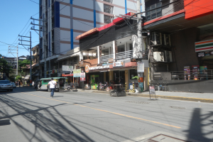 Eine Stadtstraße mit Autos und Fußgängern, hohe Gebäude mit Fenstern, Strommasten mit Drähten, Schilder, Bäume und einen klaren blauen Himmel.