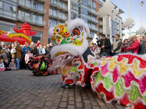 Ein lebendiges chinesisches Neujahrsfest in Amsterdam mit einer Löwendance und einer Menge Schaulustiger, einige fotografieren das Ereignis, vor einer Kulisse aus Gebäuden, Laternenmasten und einem klaren blauen Himmel.