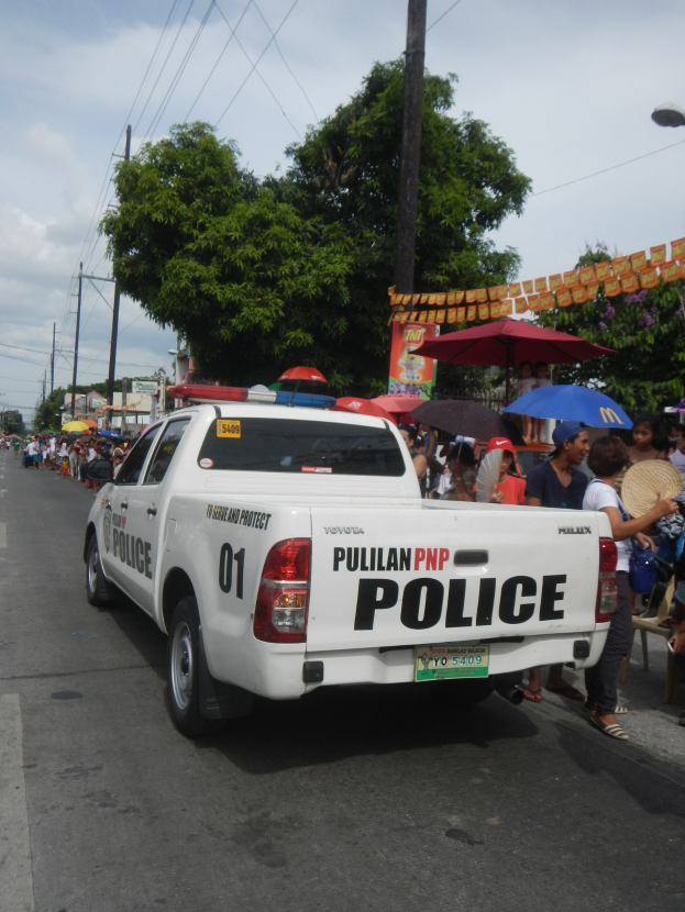Polizeifahrzeug fährt die Straße entlang, Menschen mit Schirmen auf dem Gehweg, Bäume und Gebäude im Hintergrund bei klarem blauem Himmel.