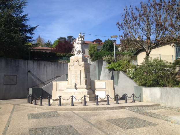Eine Bronzestatue eines Mannes steht auf einem Sockel in einem Park, umgeben von Bäumen, einem Metallzaun und einem bewölkten Himmel, vermutlich ein Mahnmal für die Opfer der Pariser Anschläge.