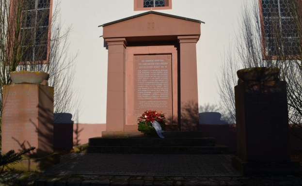 Ein Gebäude mit Fenstern, ein Denkmal mit Text, ein Blumenstrauß auf dem Boden, Topfpflanzen, eine Baumgruppe und ein bewölkter Himmel, mit einem Grab und einem Kränzchen davor.