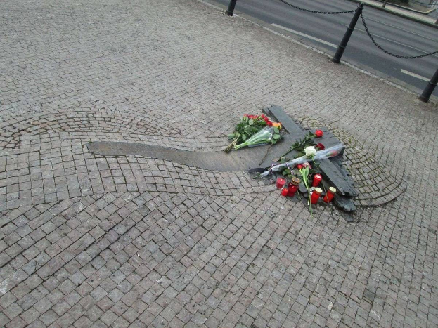Gedenken mit Blumen, Kerzen und verstreuten Huldigungen am Straßenrand, umgeben von Bäumen, Gebäuden und Fahrzeugen.