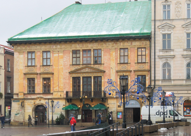 Ein großes Gebäude mit einem grünen Dach in einer Stadt, umgeben von Straßenmasten, Straßenlaternen, Fahrzeugen, Fußgängern, Geländern, Bäumen und einem klaren blauen Himmel.