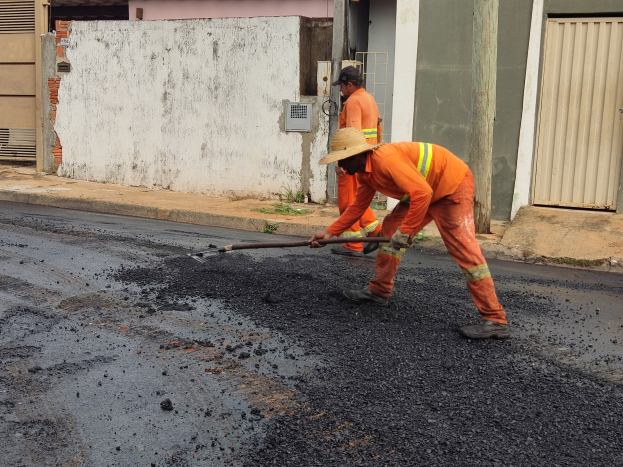 Zwei Männer in orangefarbenen Overalls und Hüten bei der Arbeit auf einer Straße, einer hält ein Objekt, mit Gebäuden und Rollläden im Hintergrund.