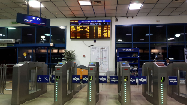 Ein Mann steht vor einer Metalldrehsperre am Eingang eines Flughafens, mit Metalldetektoren, Informationsschildern, einem Topf mit einer Pflanze, einer Wandtafel, Deckenleuchten und einem Anzeigebildschirm im Hintergrund.