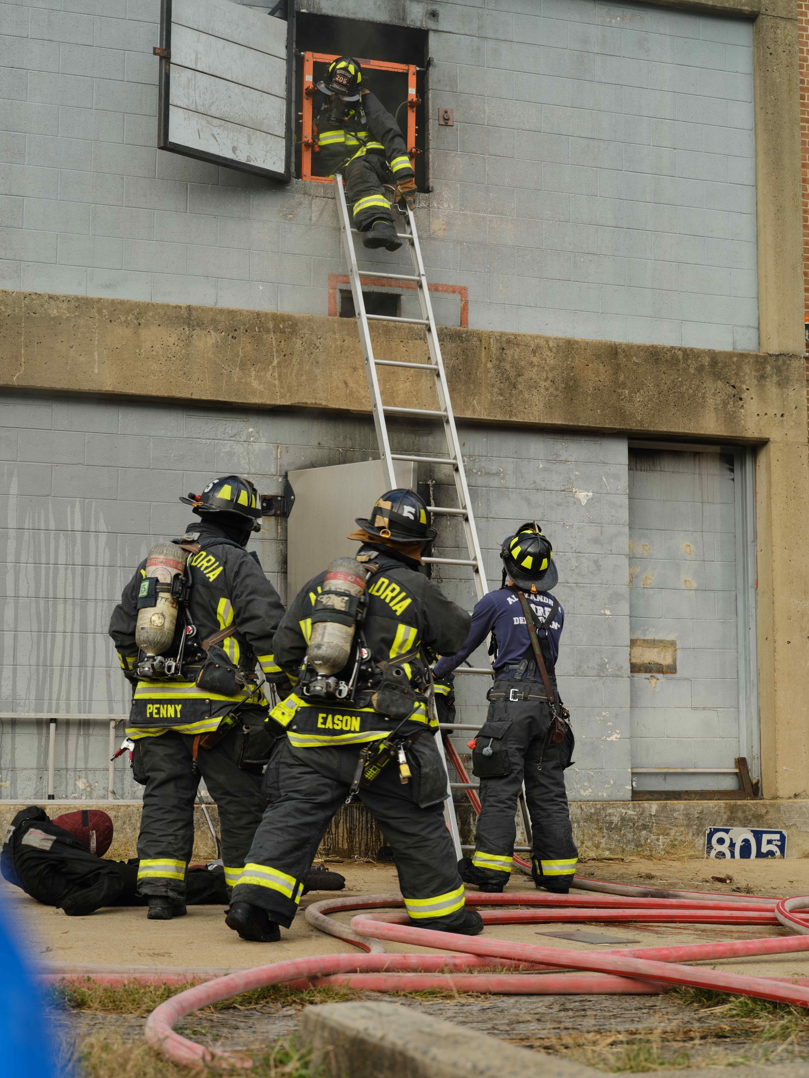 Feuerwehrleute in Helmen und Ausrüstung, die an einem Gebäude eine Leiter hochsteigen, mit Rohren und Gras unten und einer Person in der Nähe.