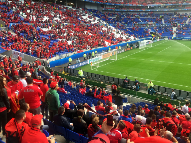 Ein volles Stadion mit Zuschauern, einige sitzen und einige stehen, die ein Fußballspiel beobachten, mit sichtbaren Torpfosten, Bannern und Deckenleuchten unter einer Dachkonstruktion.
