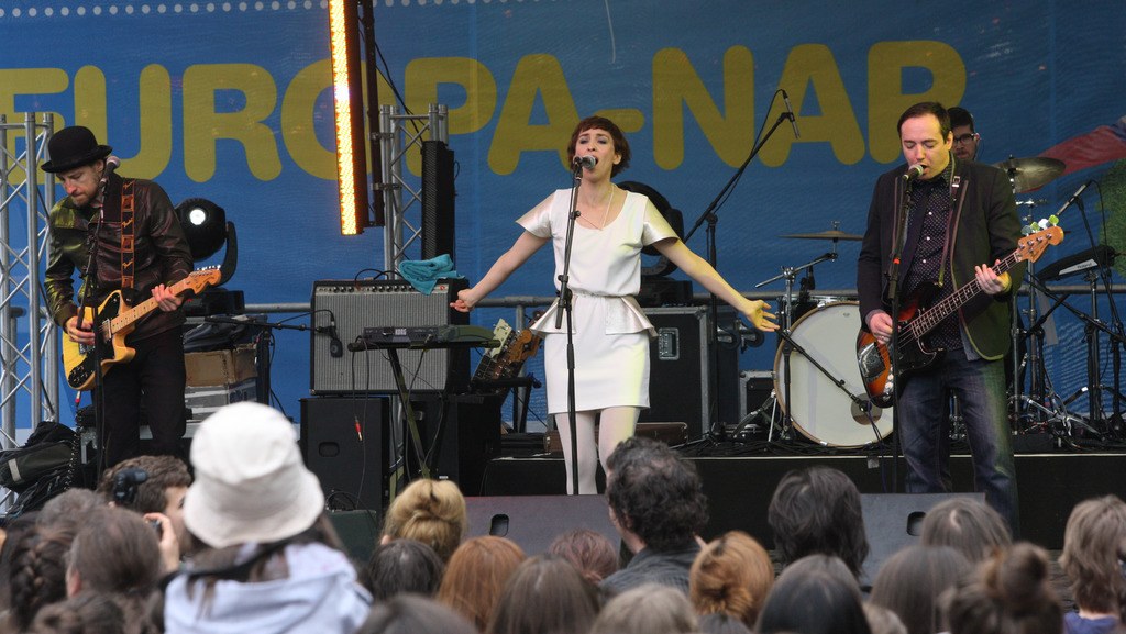 Vier Personen auf der Bühne: Eine Frau in einem weißen Kleid singt, flankiert von zwei Männern, die Gitarren spielen, mit einer Menge unten und Lautsprechern an der Wand dahinter.