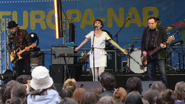 Vier Personen auf der Bühne: Eine Frau in einem weißen Kleid singt, flankiert von zwei Männern, die Gitarren spielen, mit einer Menge unten und Lautsprechern an der Wand dahinter.