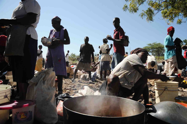 Eine Gruppe von Menschen, die in l├Ąndlicher Gegend im Freien Essen zubereiten, mit einem gro├čen Topf, Eimern und Abdeckungen auf dem Boden, unter einem klaren blauen Himmel.