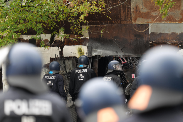 Eine Gruppe von Polizisten in schwarzen Uniformen und Helmen vor einem Gebäude, das von einem Maschendrahtzaun umgeben ist, mit Bäumen im Hintergrund.