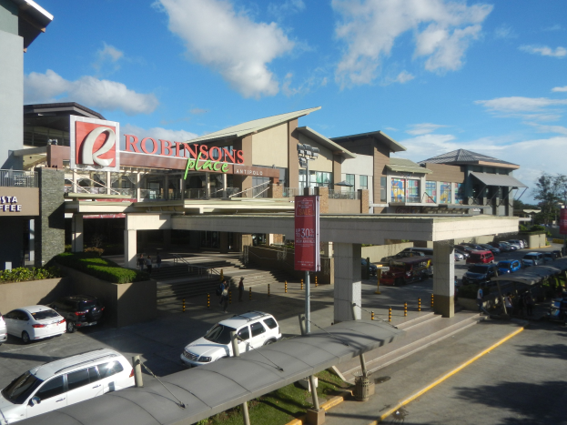 Ein belebter Einkaufszentrum mit Gebäuden, Geschäften, Fahrzeugen, Fußgängern, Straßenmöblierung, Grünflächen und einem bewölkten Himmel.