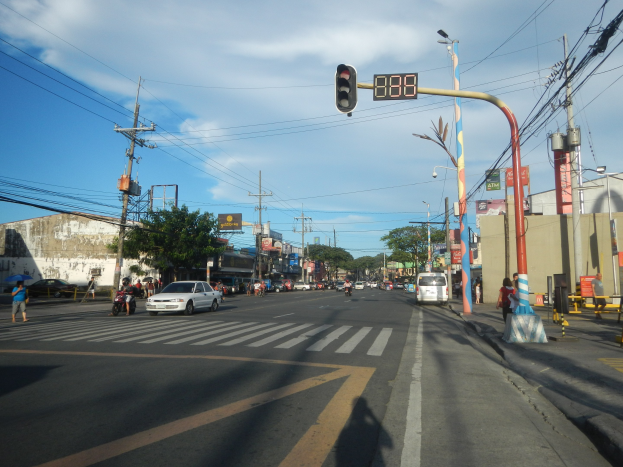 Eine Straßenkreuzung in einer Stadt mit einer Ampel, Fahrzeugen, Fußgängern, Strommasten, Laternen, Bäumen, Gebäuden und Verkehrszeichen unter einem bewölkten Himmel.