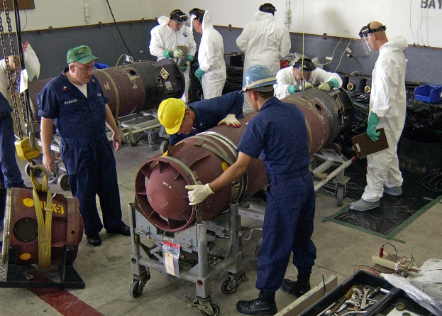 Männer in Helmen und Handschuhen bei der Entschärfung einer großen Bombe in einer Fabrik mit Maschinen und Wandtext.