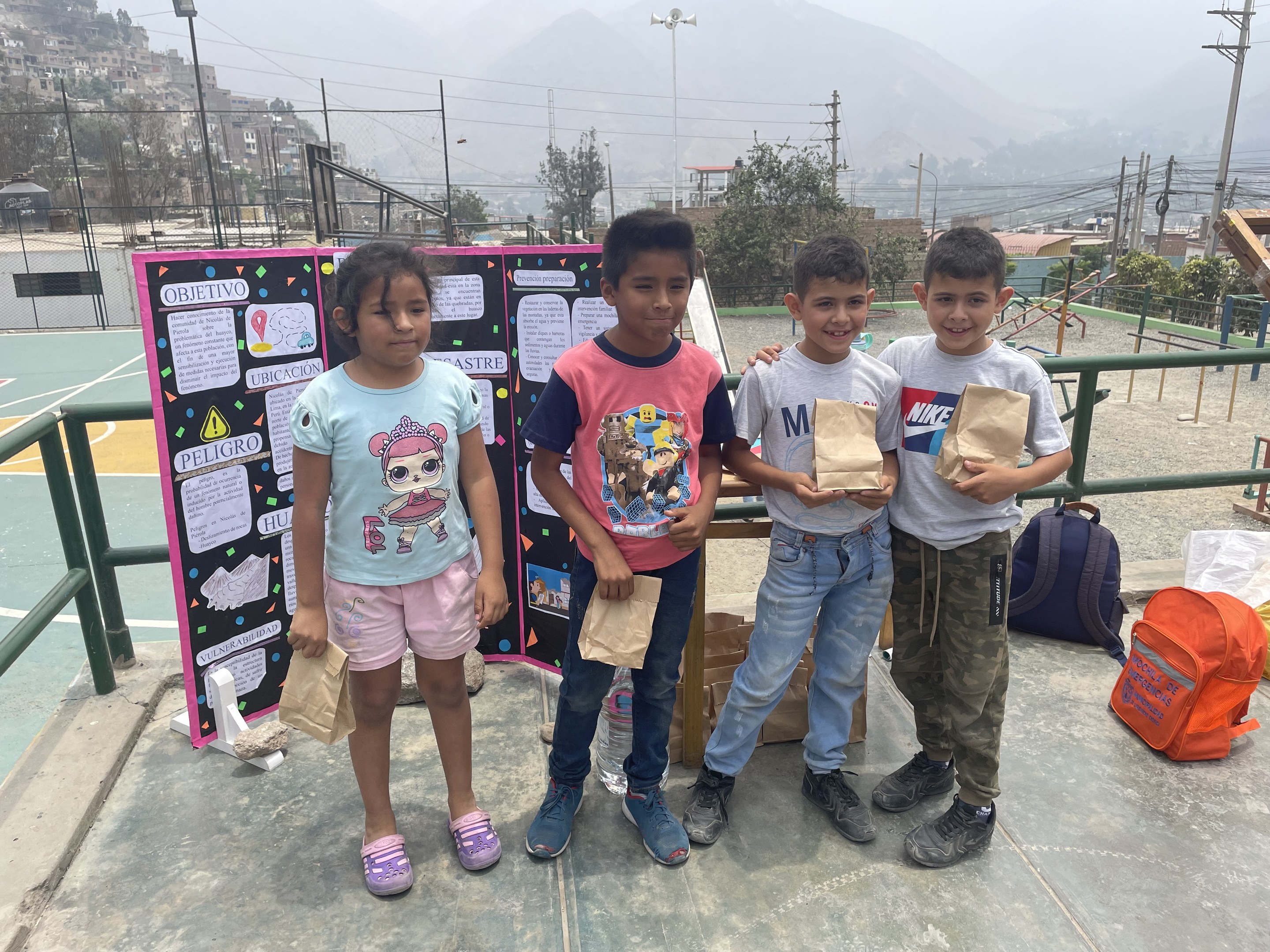 Kinder auf einem Basketballfeld mit Papierbeuteln in den Händen, zusätzliche Beutel und Gegenstände auf dem Boden, eine Tafel mit Text links und ein Hintergrund mit Zäunen, Pfosten, Straßenlaternen, Bäumen, Gebäuden, Hügeln und Himmel.