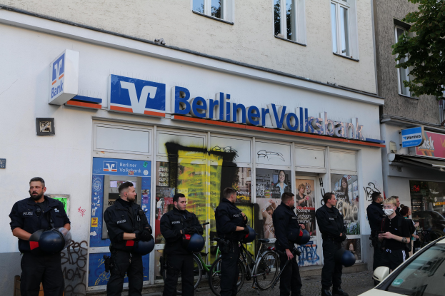 Polizeibeamte in Uniform vor einer Berliner Volksbank-Filiale, einige mit Helmen in den Händen, daneben ein geparktes Auto und ein Baum; im Hintergrund sind Gebäude mit Fenstern und Schildern zu sehen.