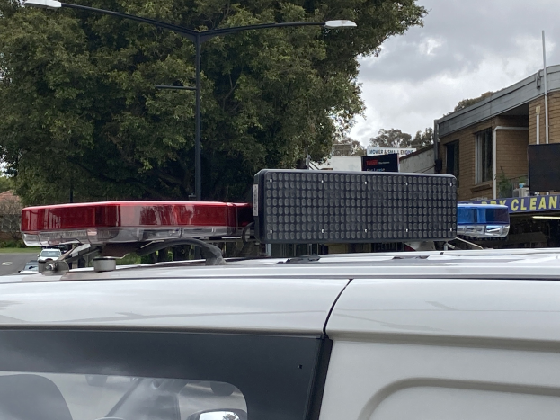 Polizeiauto mit Einsatzlichtern auf einer Straße neben einer Schule geparkt, umgeben von Gebäuden, Bäumen und Strommasten.