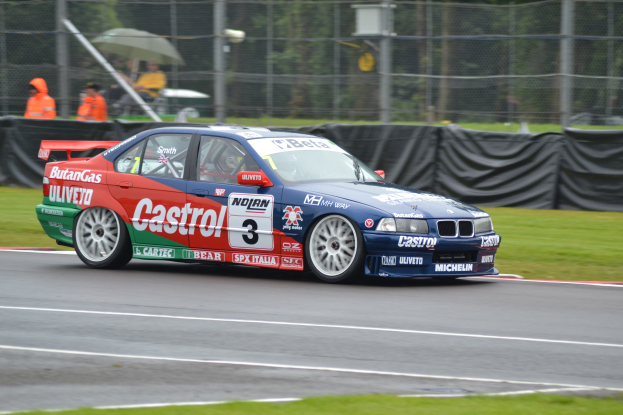 Ein Auto auf einer grasumgebenen Rennstrecke mit Text und Zahlen, mit Menschen in orangefarbenen Jacken und Schirmen hinter Maschendrahtzaun und Bäumen im Hintergrund.