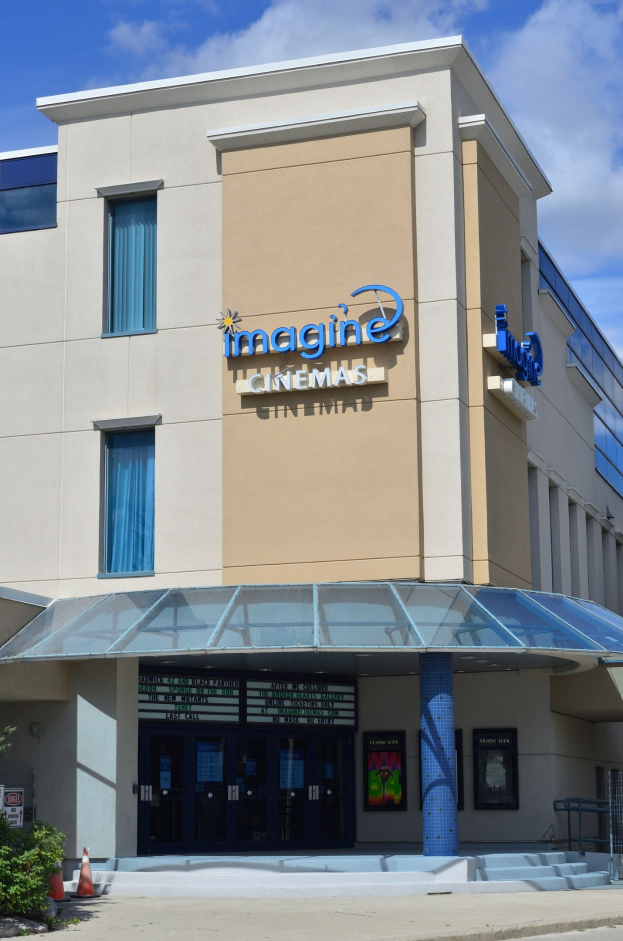 Außenansicht des Imagine Cinemas in St. Louis, Missouri, mit Glasfenstern und -türen, einer Schautafel, einem Metallzaun, Pflanzen, einem Verkehrspfahl, Bäumen und einem bewölkten Himmel.