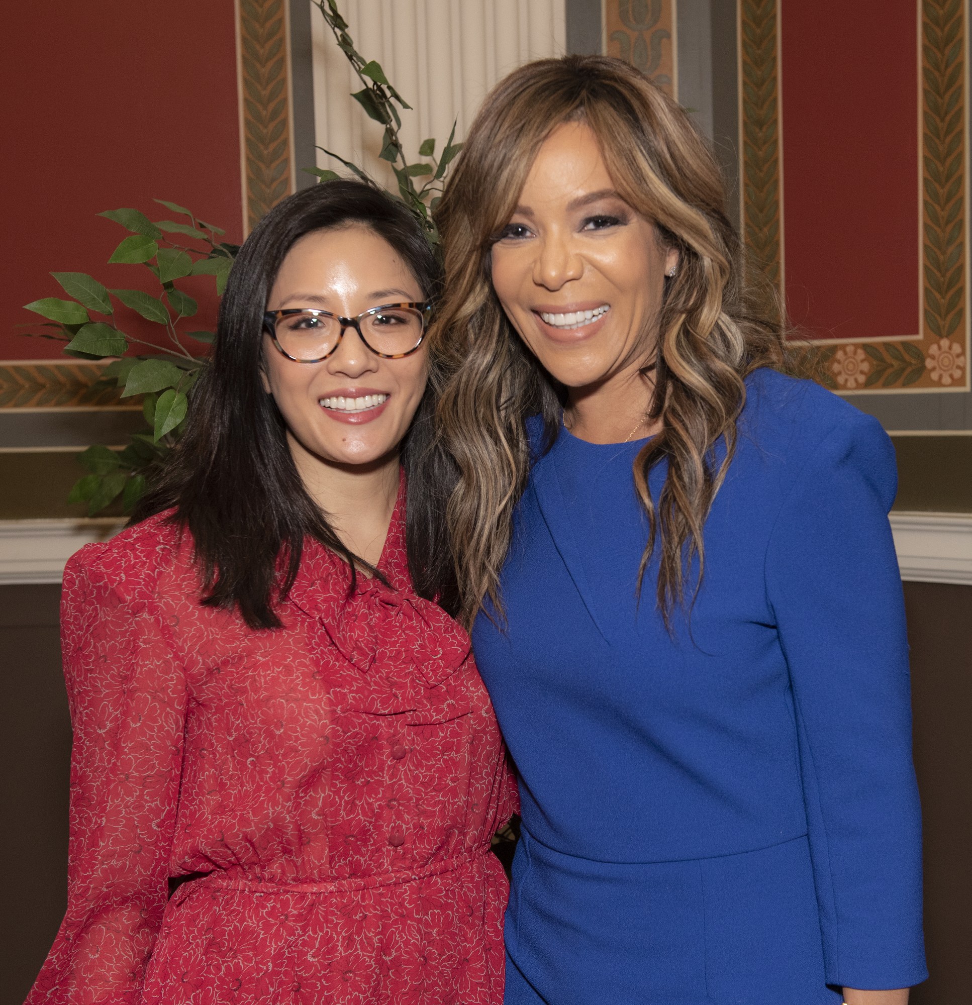 Zwei Frauen stehen nebeneinander und lächeln, wobei eine ein blaues Kleid trägt und die andere ein rotes Kleid mit Brille, vor einer Wand mit einer Pflanze und einem Vorhang auf der 2019 Women in Film Pre-Oscars Luncheon.