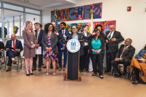 Gruppe von Menschen vor einem Rednerpult mit einem Mikrofon und einer Tafel mit der Aufschrift "Sancho City Councilman Joins City Council", mit sitzenden und stehenden Teilnehmern, Flaggen, einem Feuerlöscher und Deckenleuchten im Hintergrund.